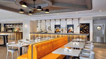 Ein modernes Restaurant mit einer zentralen orangefarbenen Essecke, weißen Tischen und hängenden Pendelleuchten.