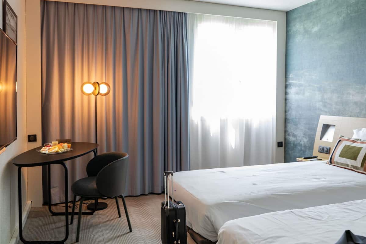 Une chambre d’hôtel avec un lit, un bureau et une chaise, une valise et une grande fenêtre avec des rideaux.