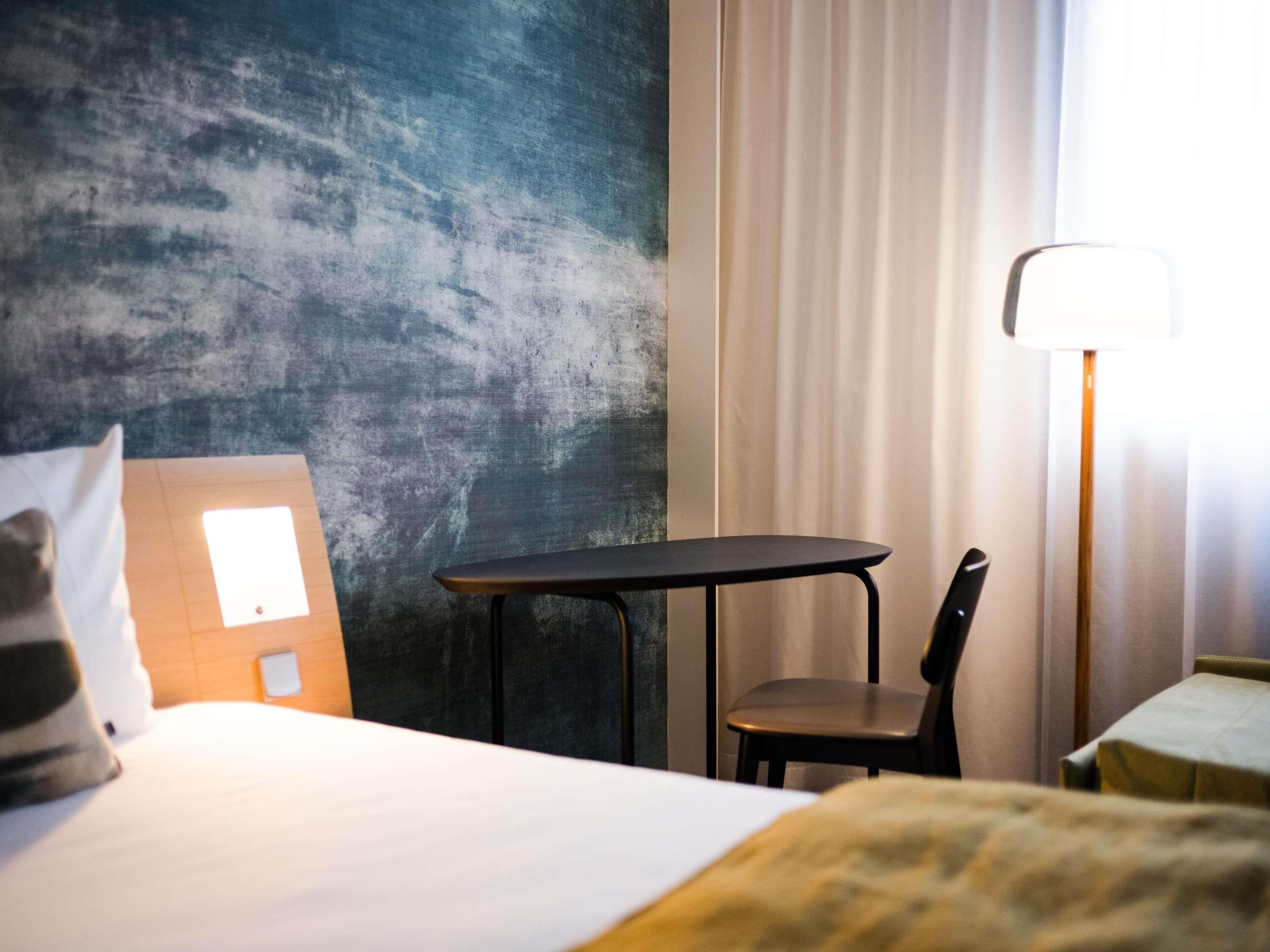 Ein Hotelzimmer mit einem Bett, einem kleinen Tisch, einem Stuhl, einer Lampe und einer Wand mit strukturierter Oberfläche.