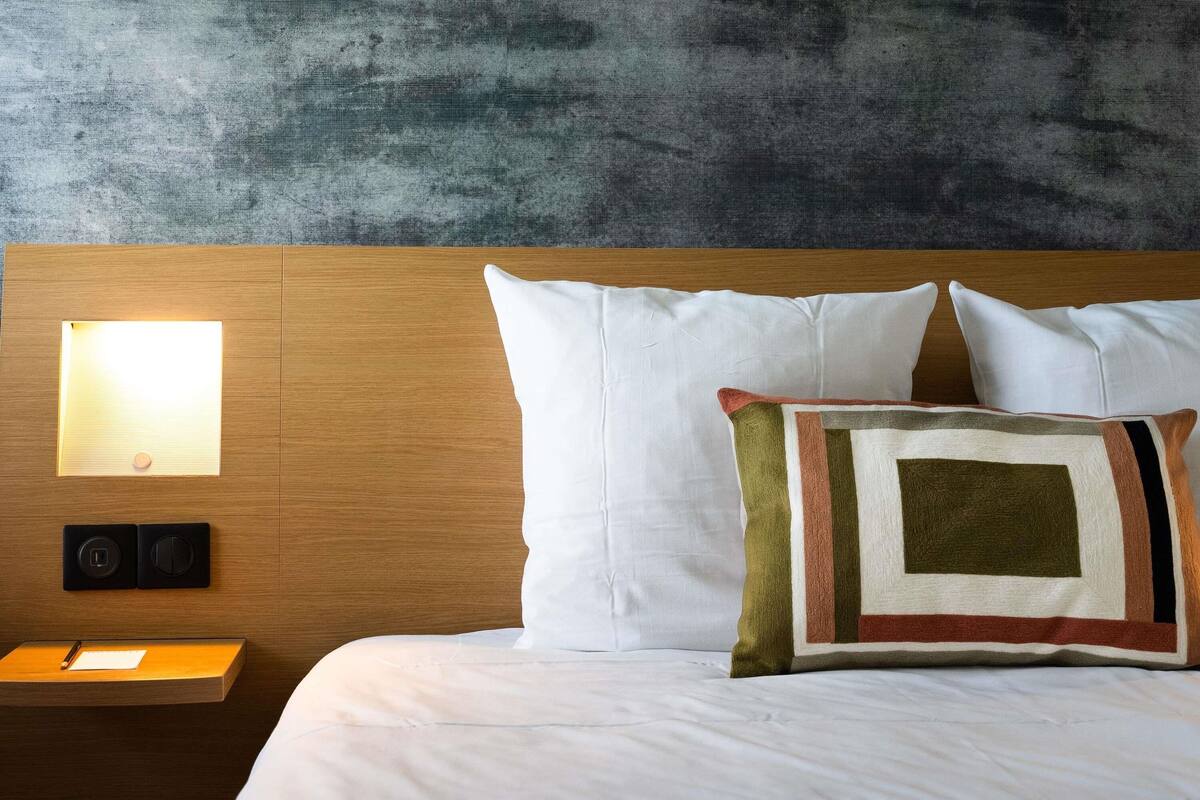 Un lit bien fait, avec du linge de lit blanc, une tête de lit en bois et un luminaire fixé au mur.