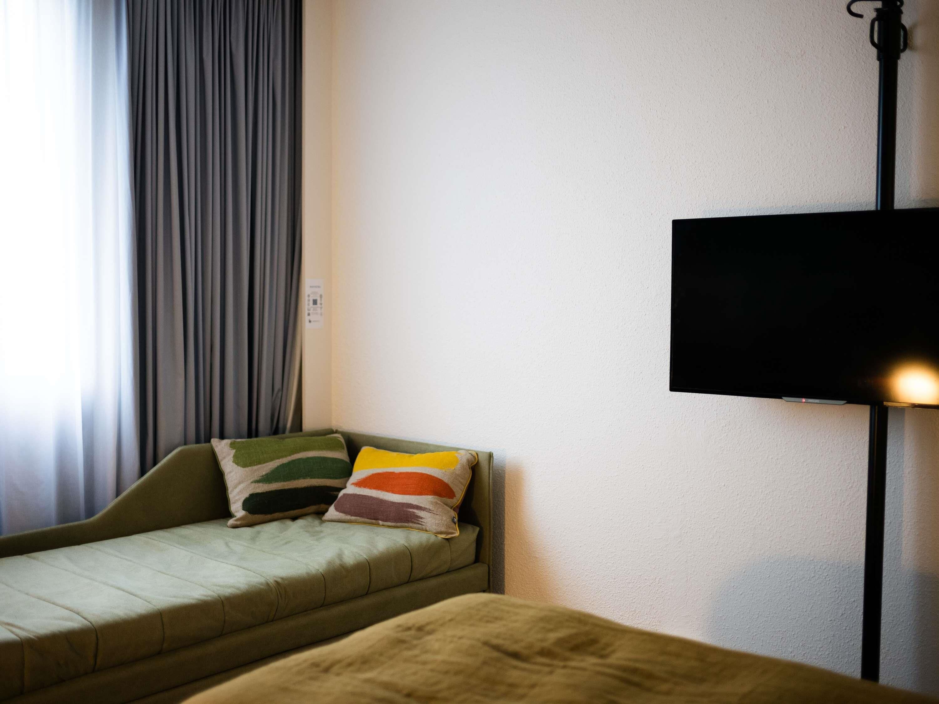 Ein Zimmer mit einer Couch, einem an der Wand befestigten Fernseher und einem Fenster mit Vorhängen.