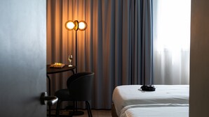 A hotel room with a bed, a desk with a chair, a lamp, and a view of the city through curtains.