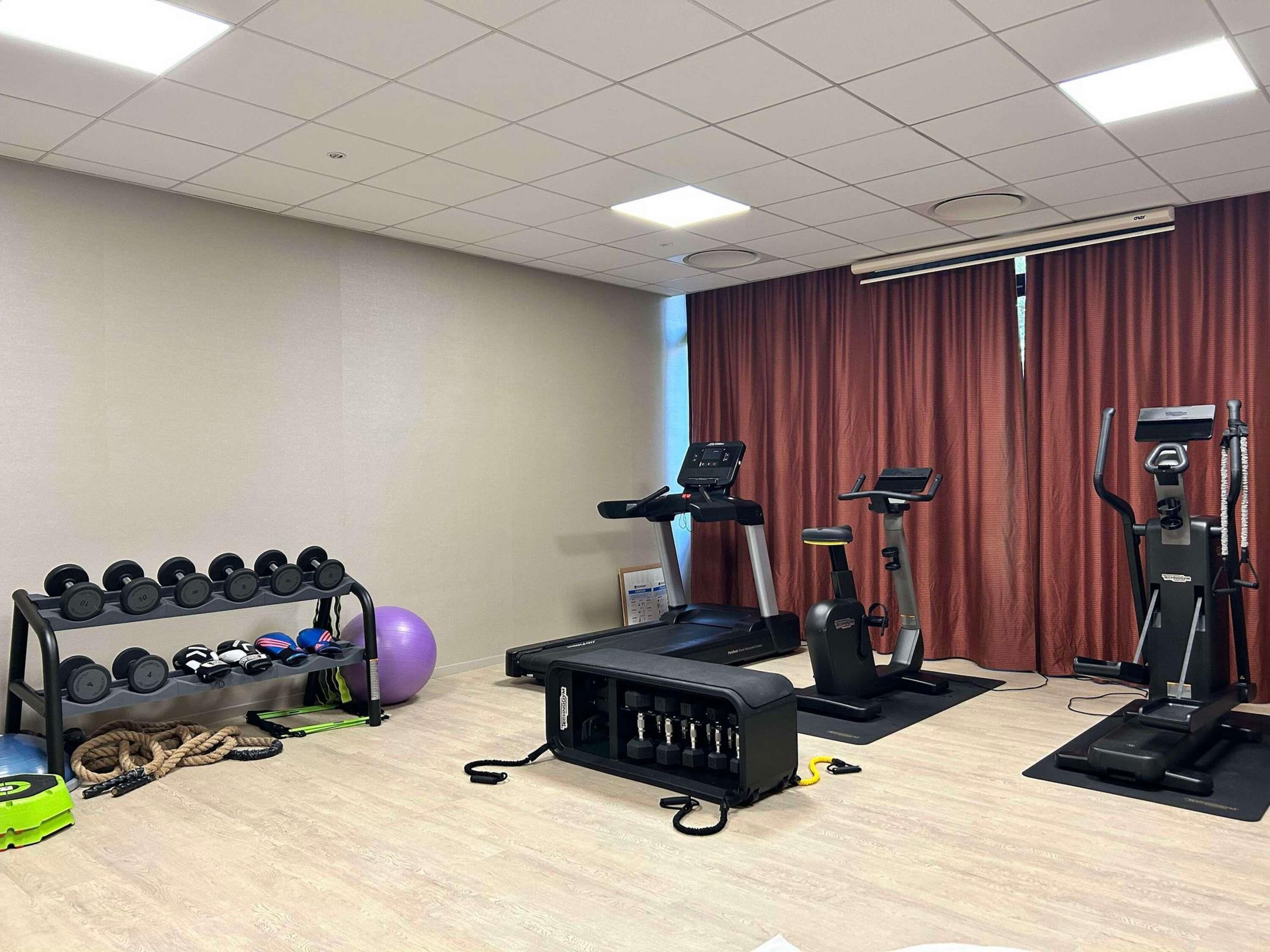 Ein Fitnessraum mit Trainingsgeräten, darunter Kurzhanteln, ein Ergometer und ein Laufband.