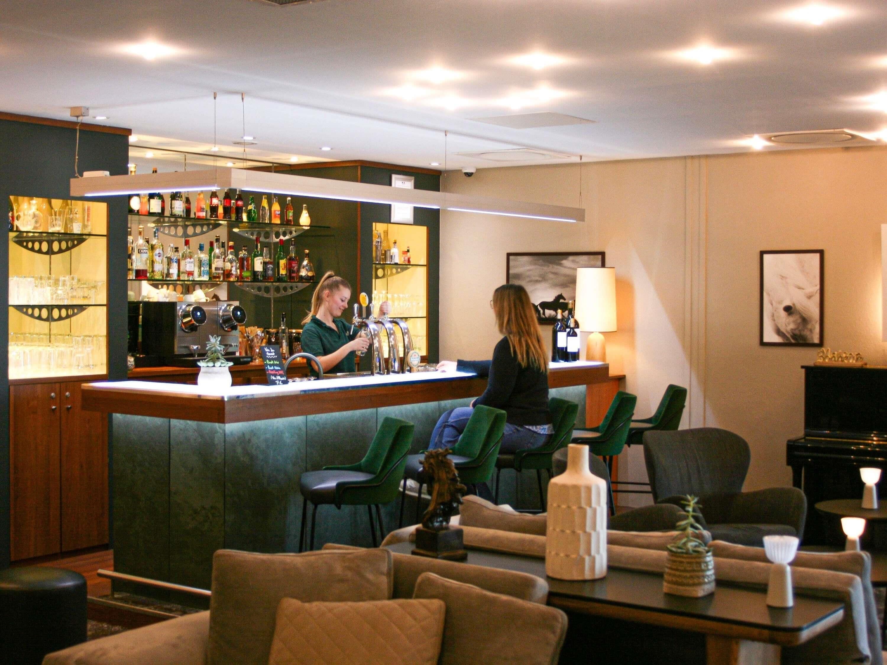 A bar area with a bartender, seating with green chairs, and a piano in the background.