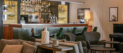 A bar area with shelves of bottles, a coffee machine, and a piano in a lounge with sofas and tables.