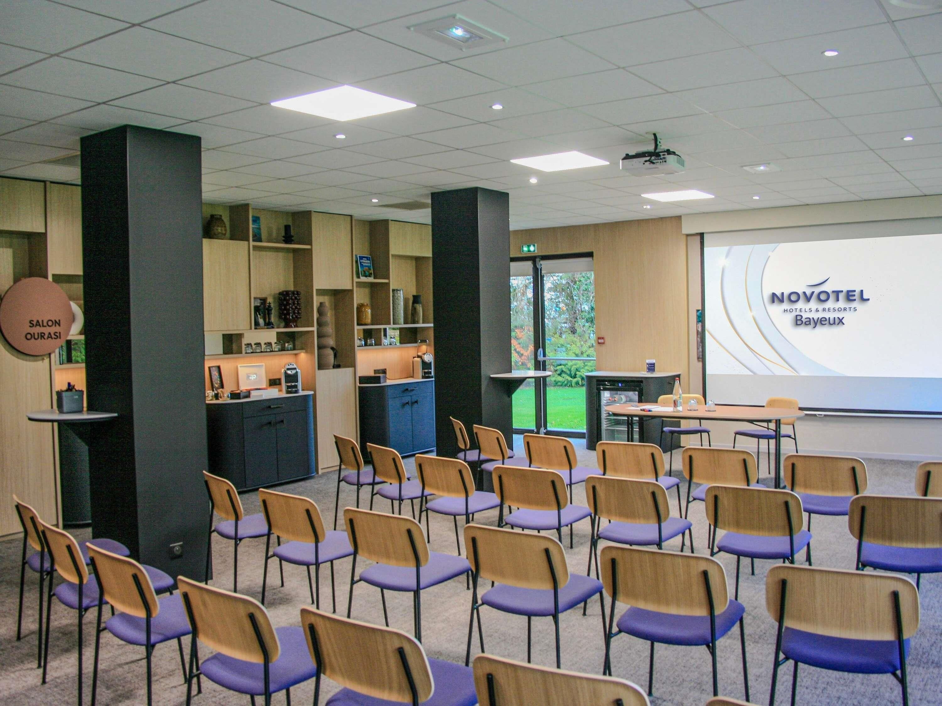 Une salle de conférence avec des rangées de chaises, un écran de présentation et une enseigne Novotel Bayeux.