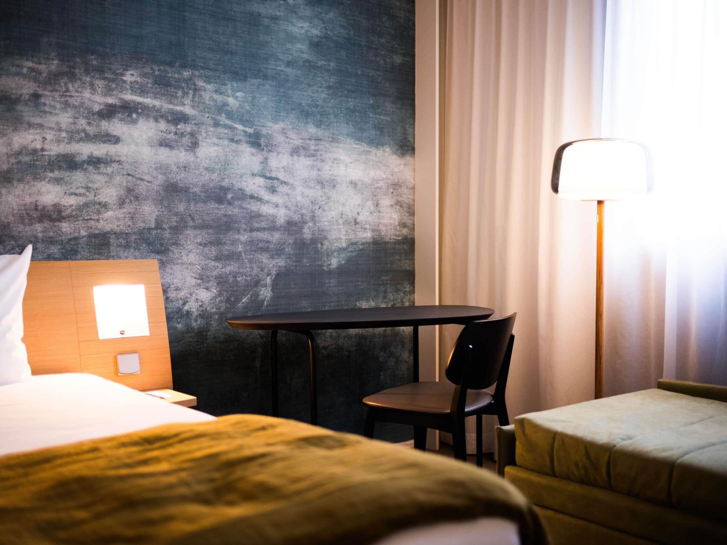 A hotel room with a bed, a small table, a chair, a sofa, and a lamp.