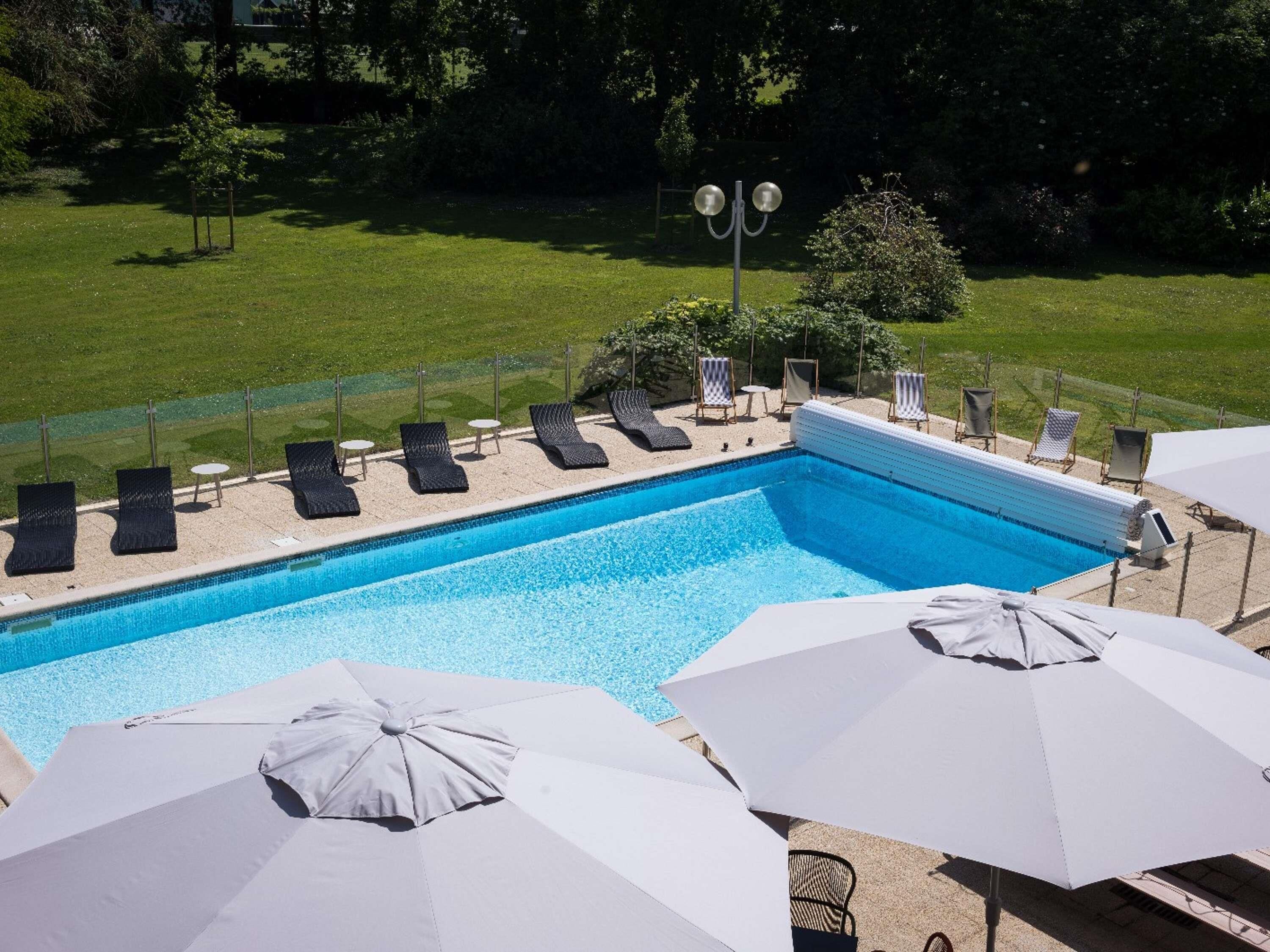 Una piscina con sillas de descanso y sombrillas, rodeada de césped y árboles.