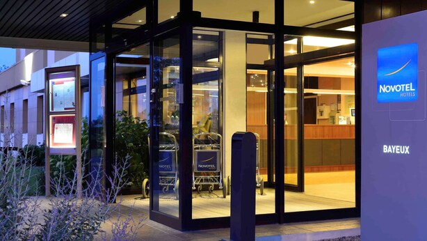 The exterior of a Novotel hotel with a glass entrance, a reception desk visible inside, and a signboard outside.