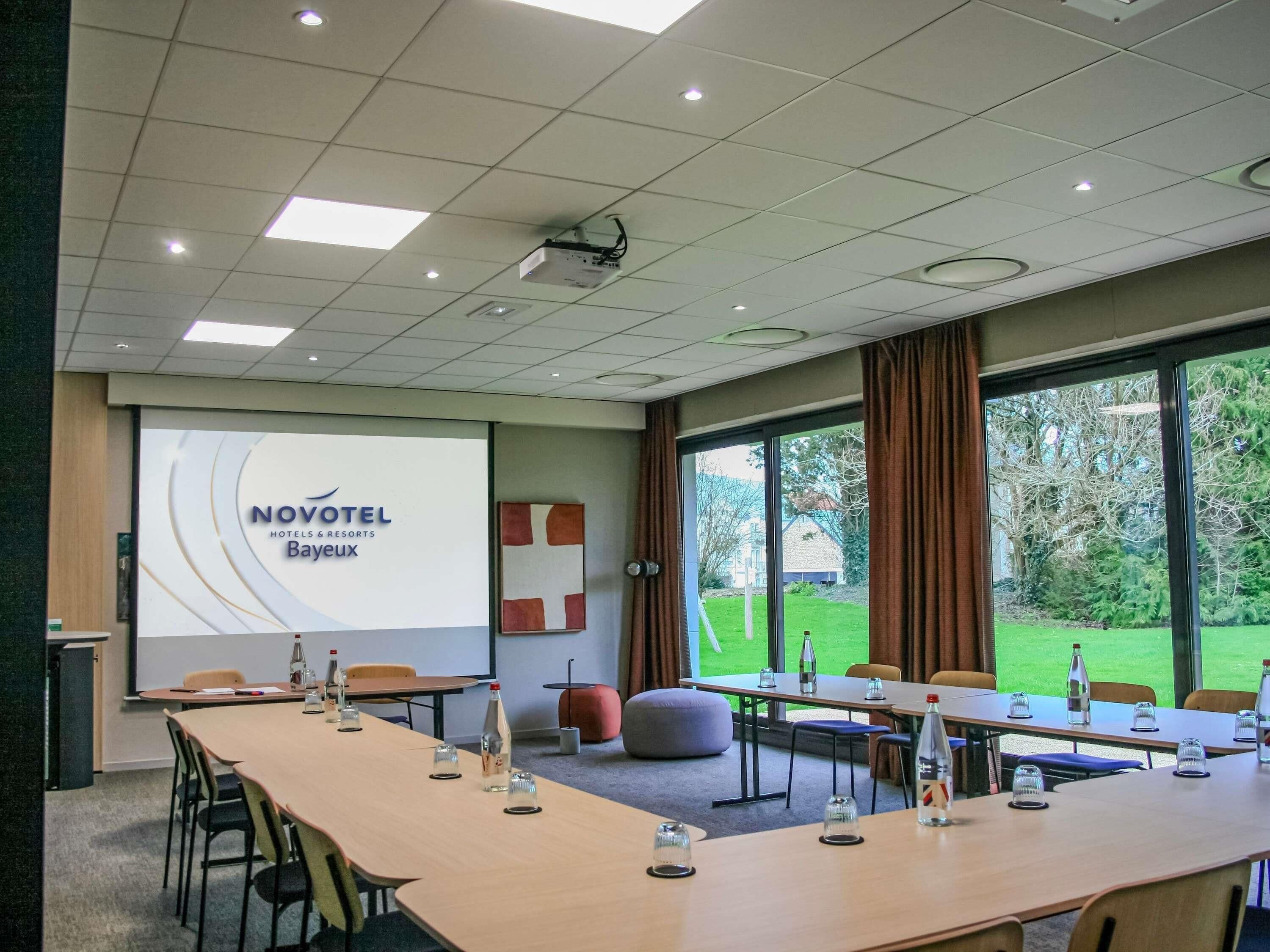 A conference room with a projector screen displaying 'NOVOTEL Hotels & Resorts Bayeux,' a long table with chairs, and large windows offering a view of greenery outside.