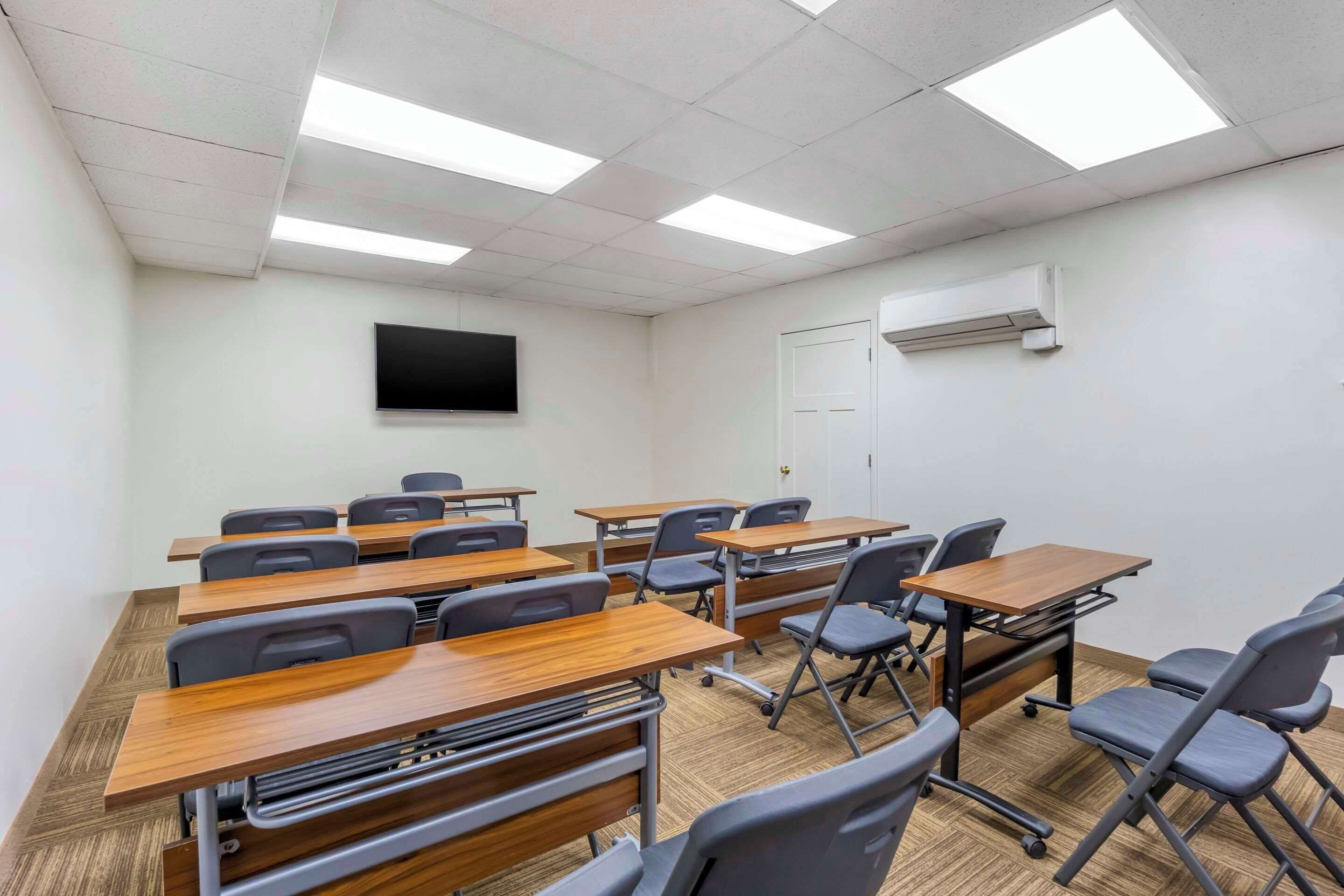 A classroom with desks and chairs, a flat-screen TV on the wall, and an air conditioning unit.