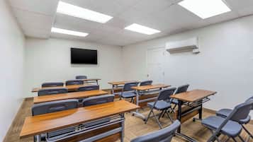 A classroom with desks and chairs, a flat-screen TV on the wall, and an air conditioning unit.