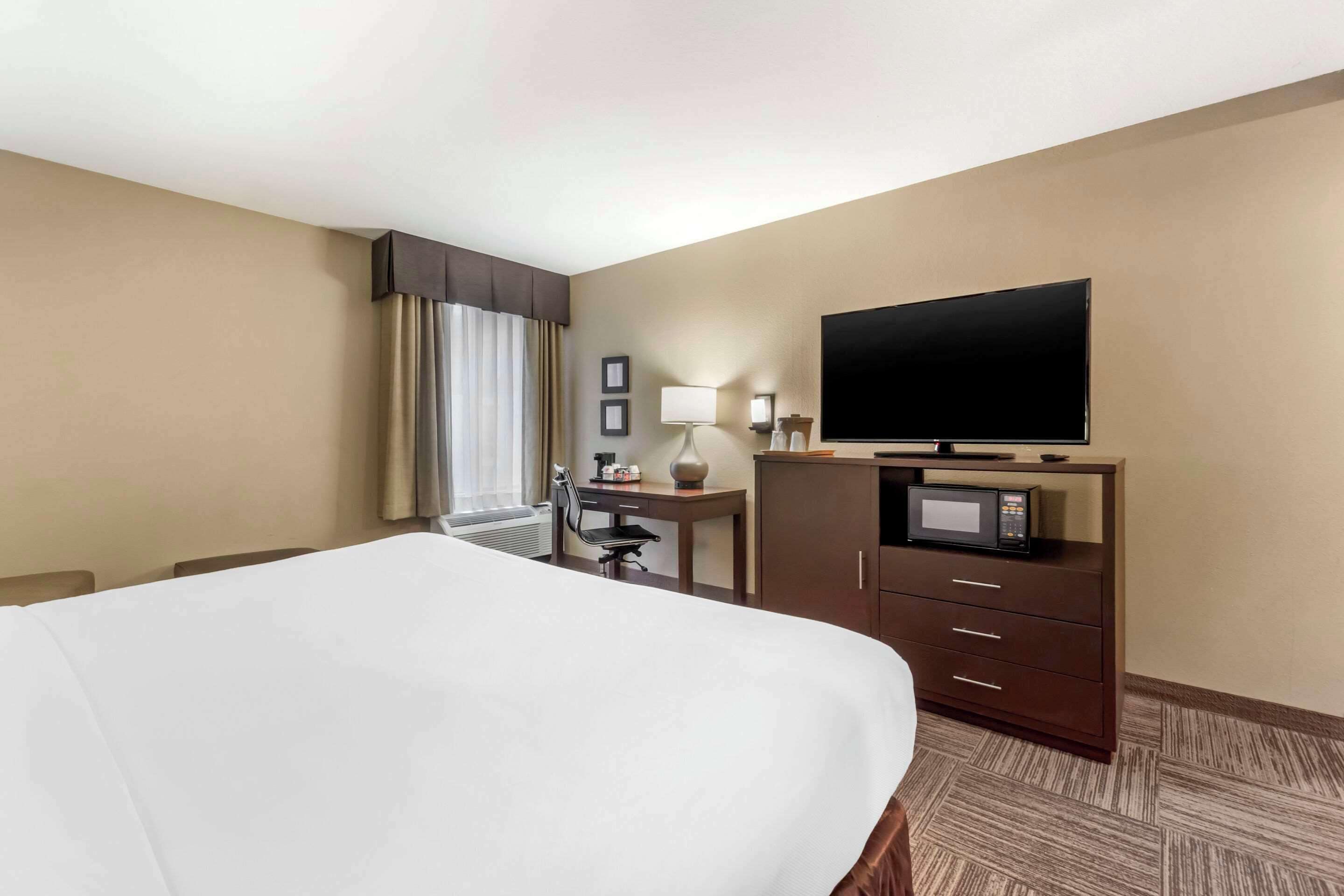 A hotel room with a large bed, a desk with a chair, a flat-screen TV, a microwave, and a window with curtains.