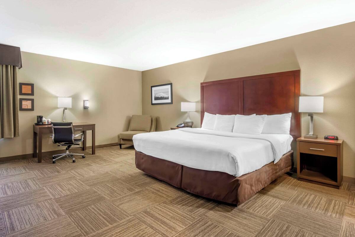A hotel room with a large bed, a desk with a chair, a small table, and a nightstand with a lamp.
