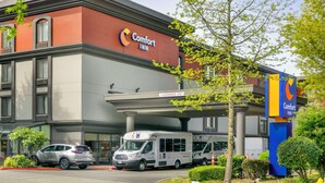 A Comfort Inn hotel with a clear sign, a covered parking area, and a parking lot with vehicles. - Comfort Inn & Suites Sea-Tac Airport (SeaTac)