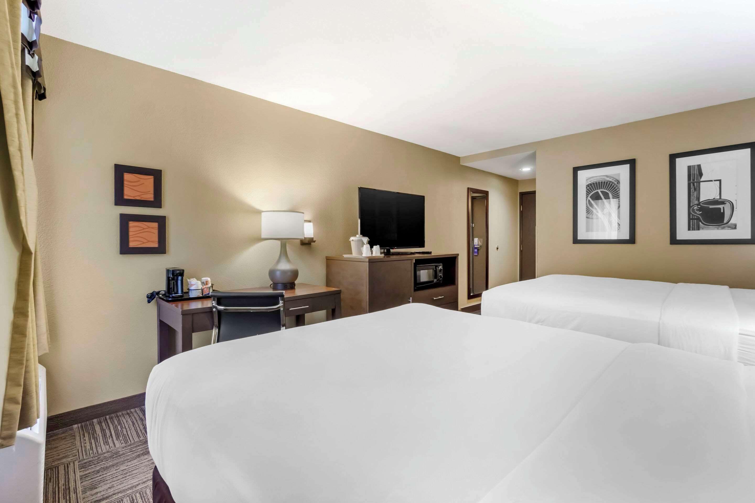 A hotel room with two beds, a desk with a lamp, a television, a microwave, and framed pictures on the wall.