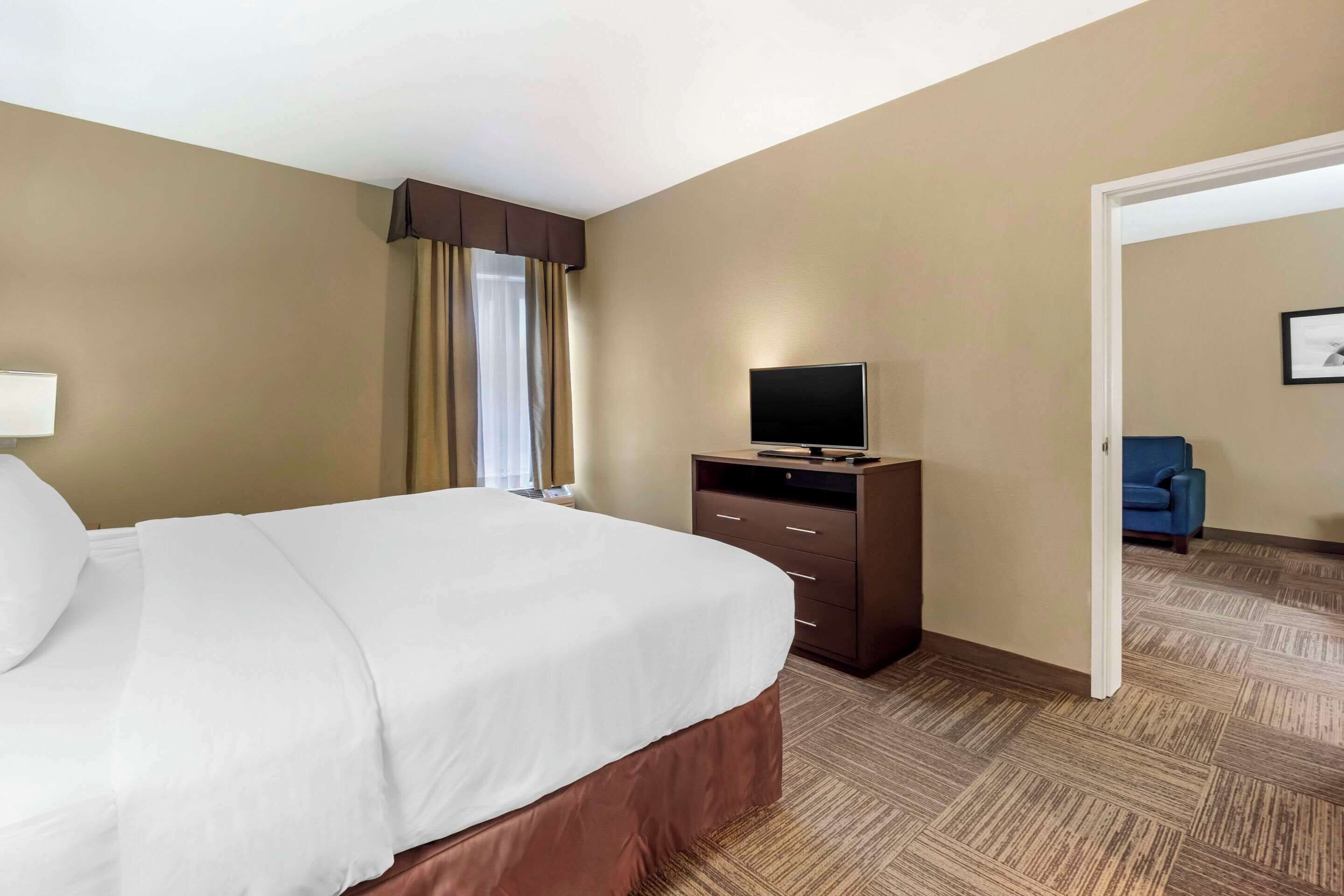 Ett hotellrum med en stor säng, en platt-TV på en byrå, ett fönster med gardiner och en blå stol i hörnet.