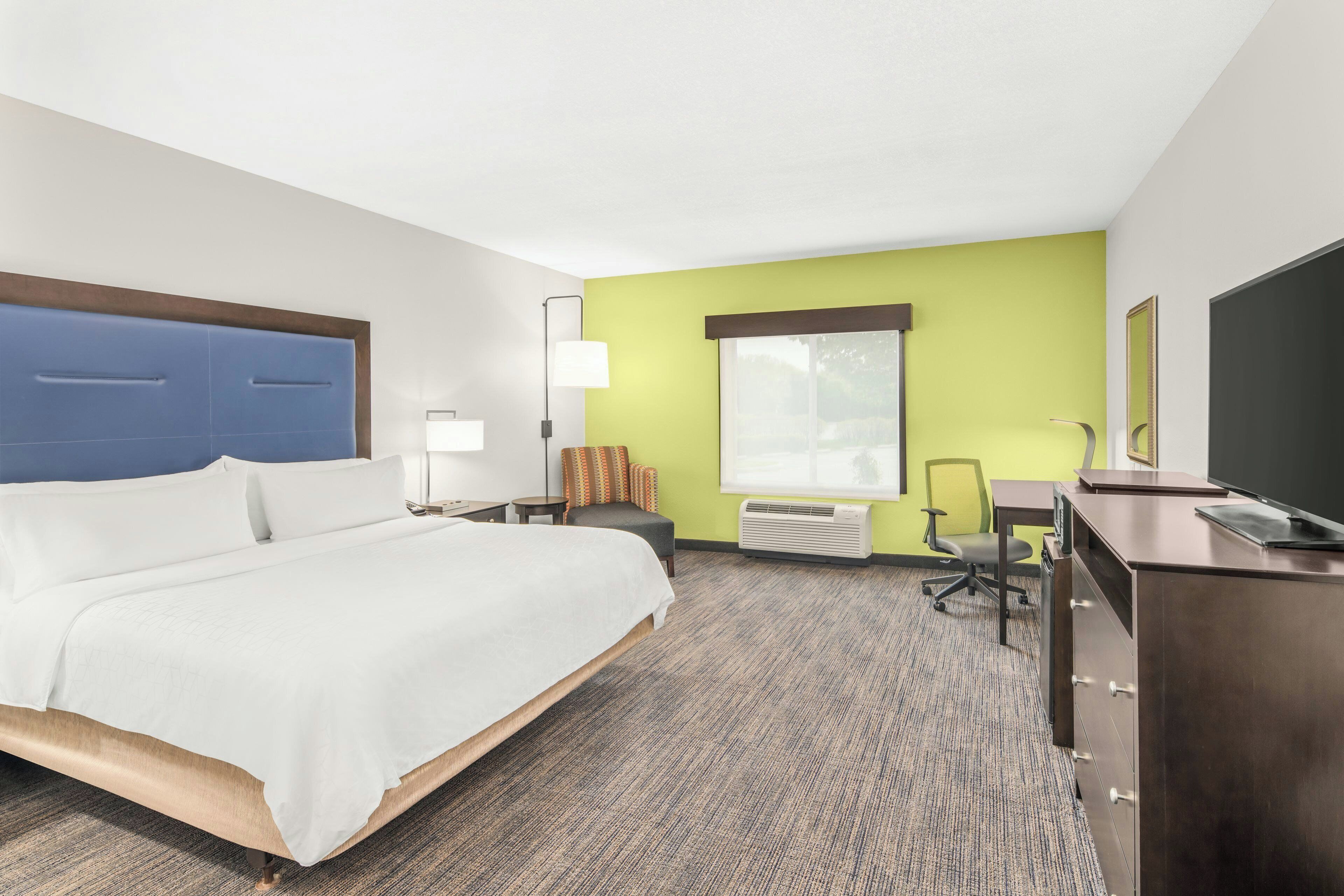 A hotel room with a bed, desk, chair, lamp, air conditioning unit, and a television.