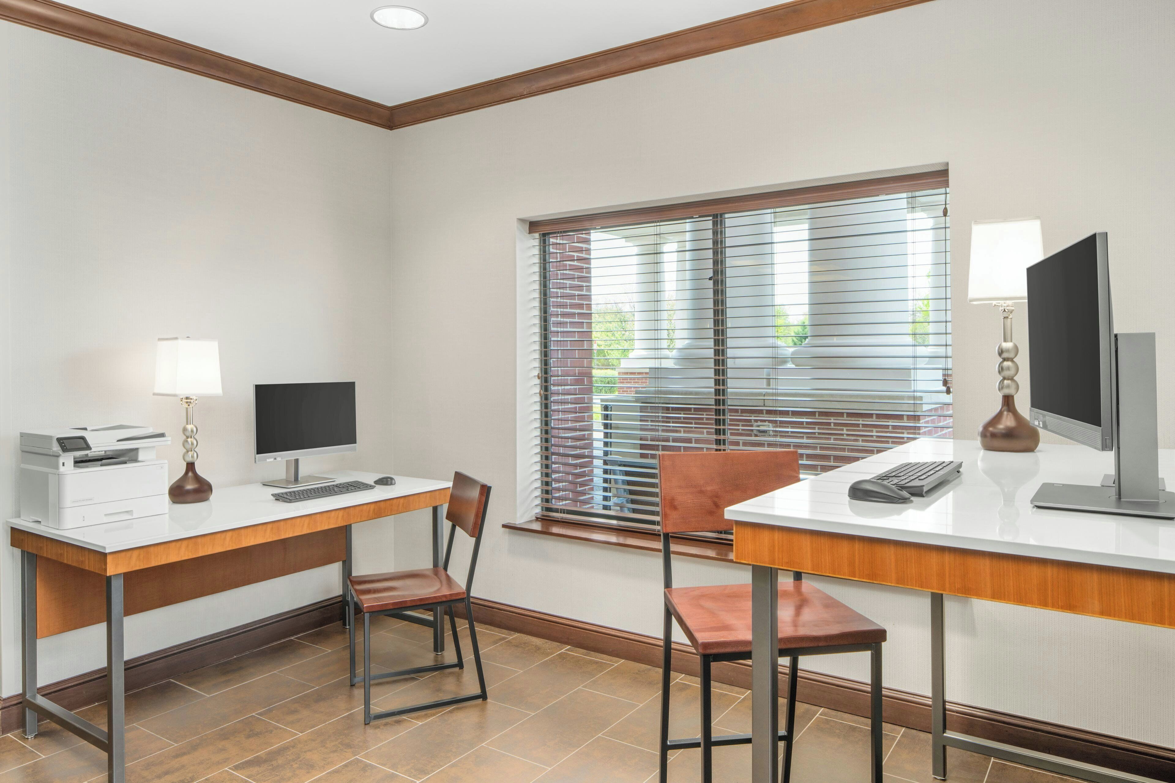 Habitación de hotel con escritorio, dos sillas, computadora, impresora y ventana con persianas.