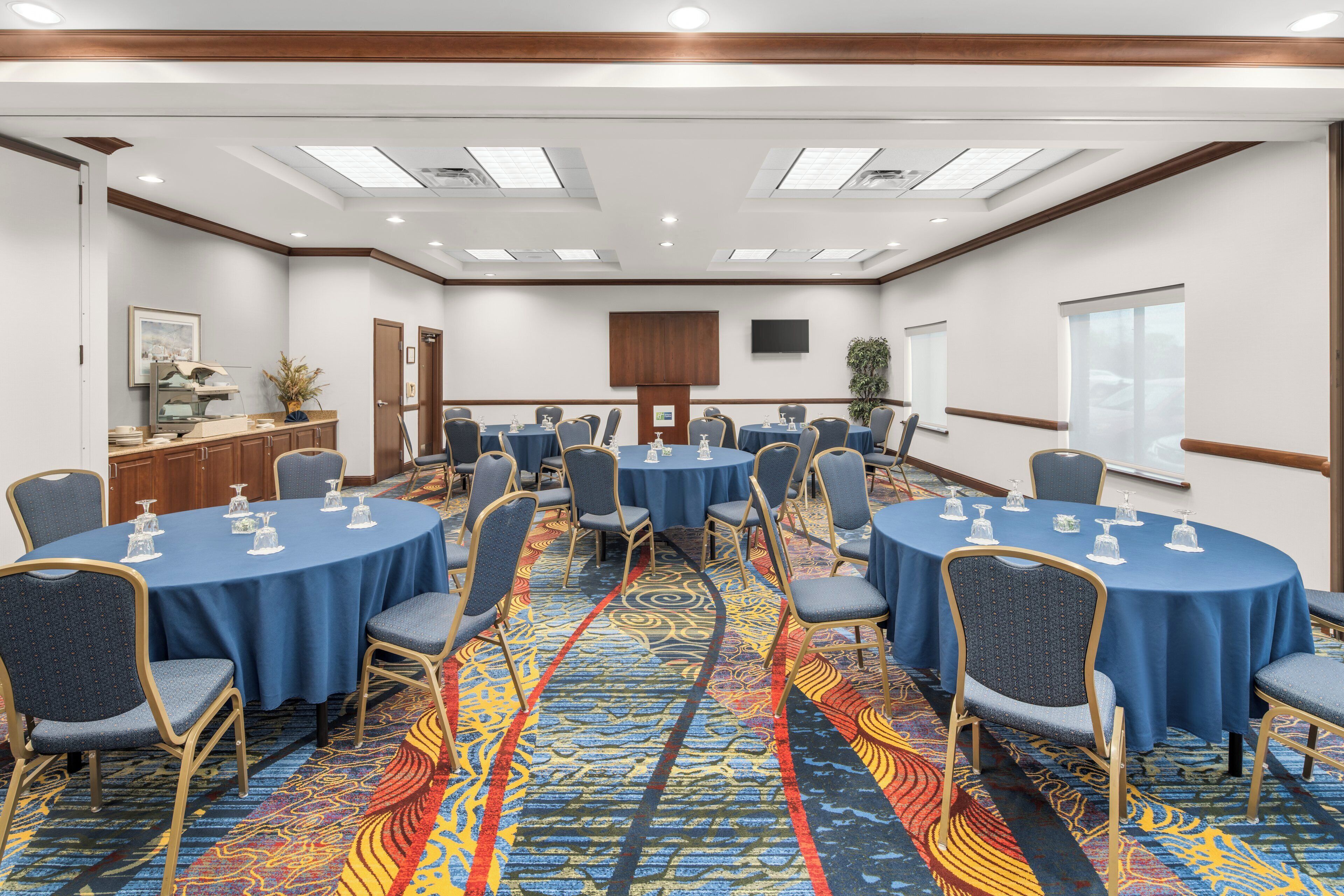 A conference room with multiple round tables, chairs, a carpeted floor, and a television on the wall.