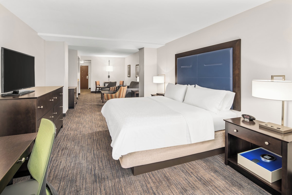 A hotel room with a large bed, a desk with a chair, a TV, and a desk lamp.