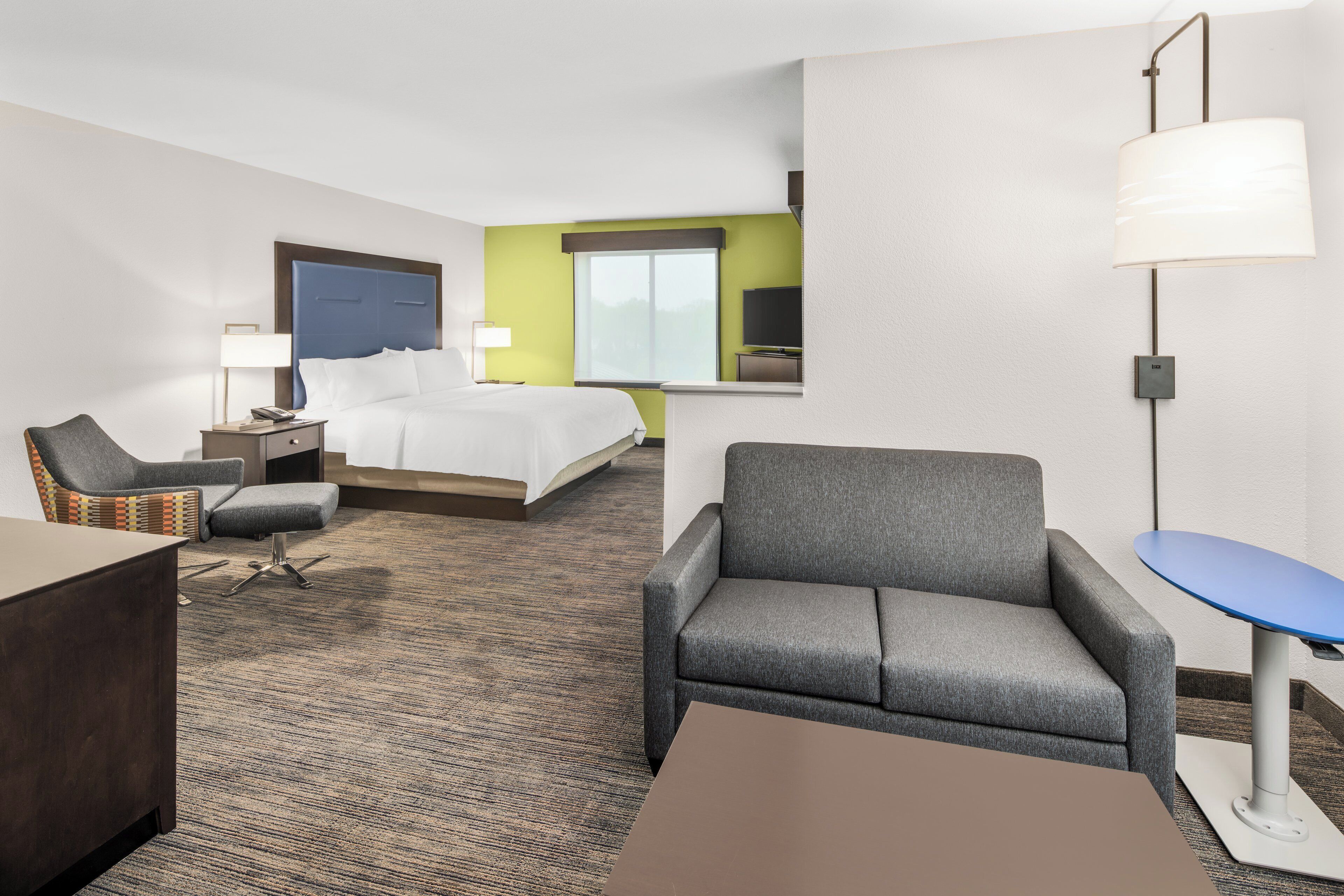 A hotel room with a bed, two armchairs, a small table, a desk, a lamp, a TV, and a window.
