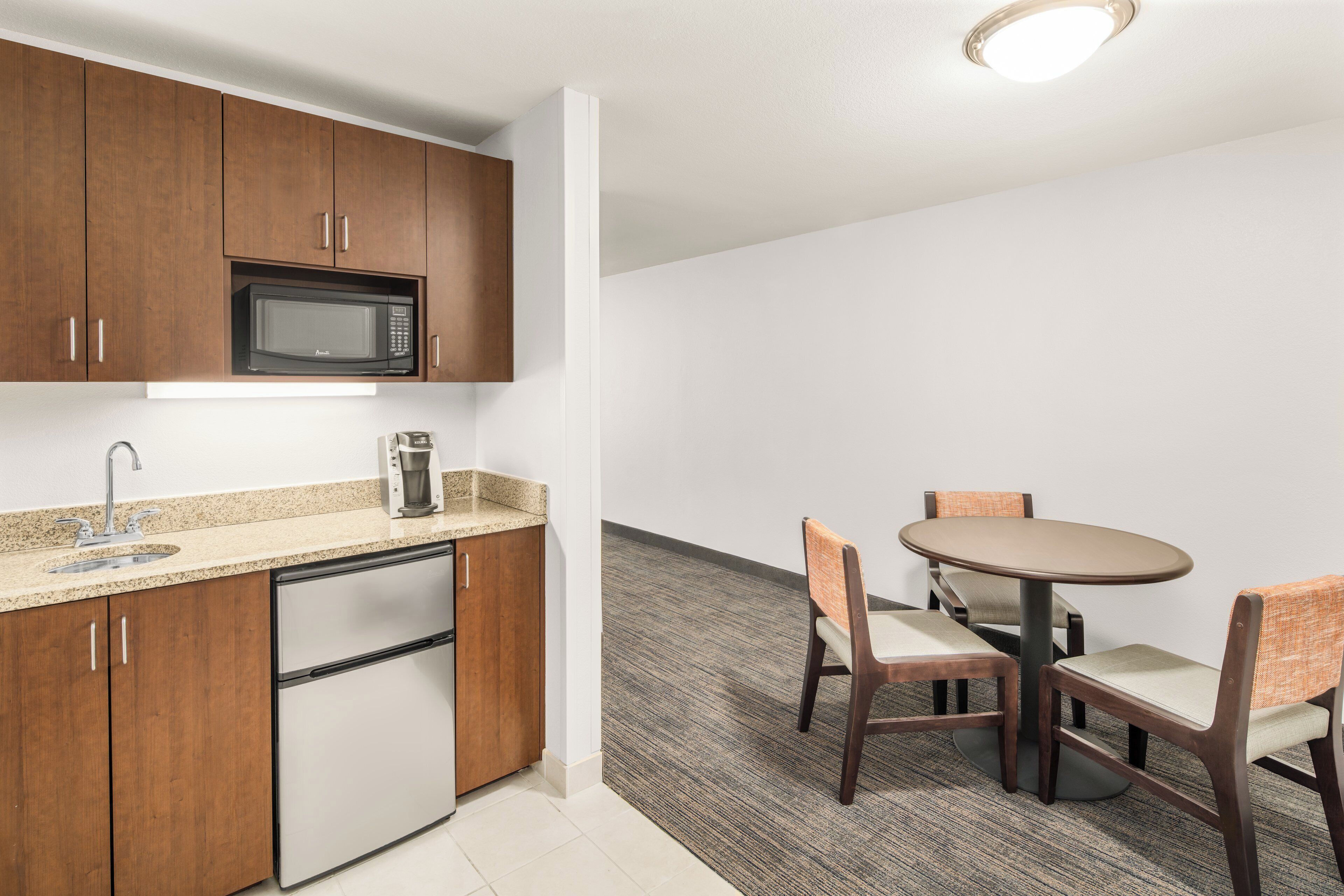 Una kitchenette compacta equipada con microondas, cafetera y lavavajillas. Cuenta con un pequeño comedor con una mesa y dos sillas.