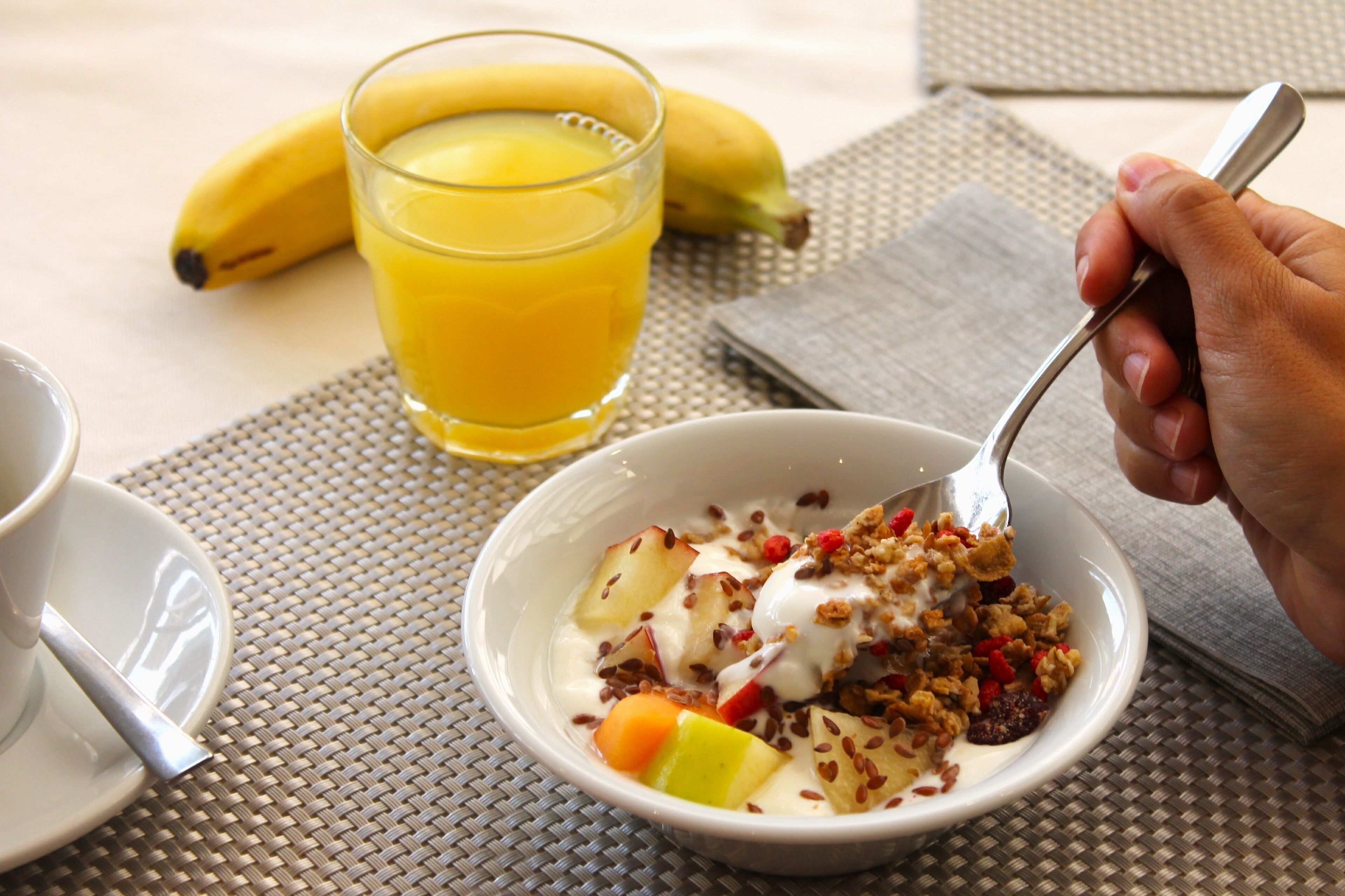 Desayuno con un tazón de cereales, yogur, fruta, un vaso de jugo de naranja y un plátano sobre la mesa.