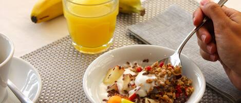 Desayuno con un tazón de cereales, yogur, fruta, un vaso de jugo de naranja y un plátano sobre la mesa.