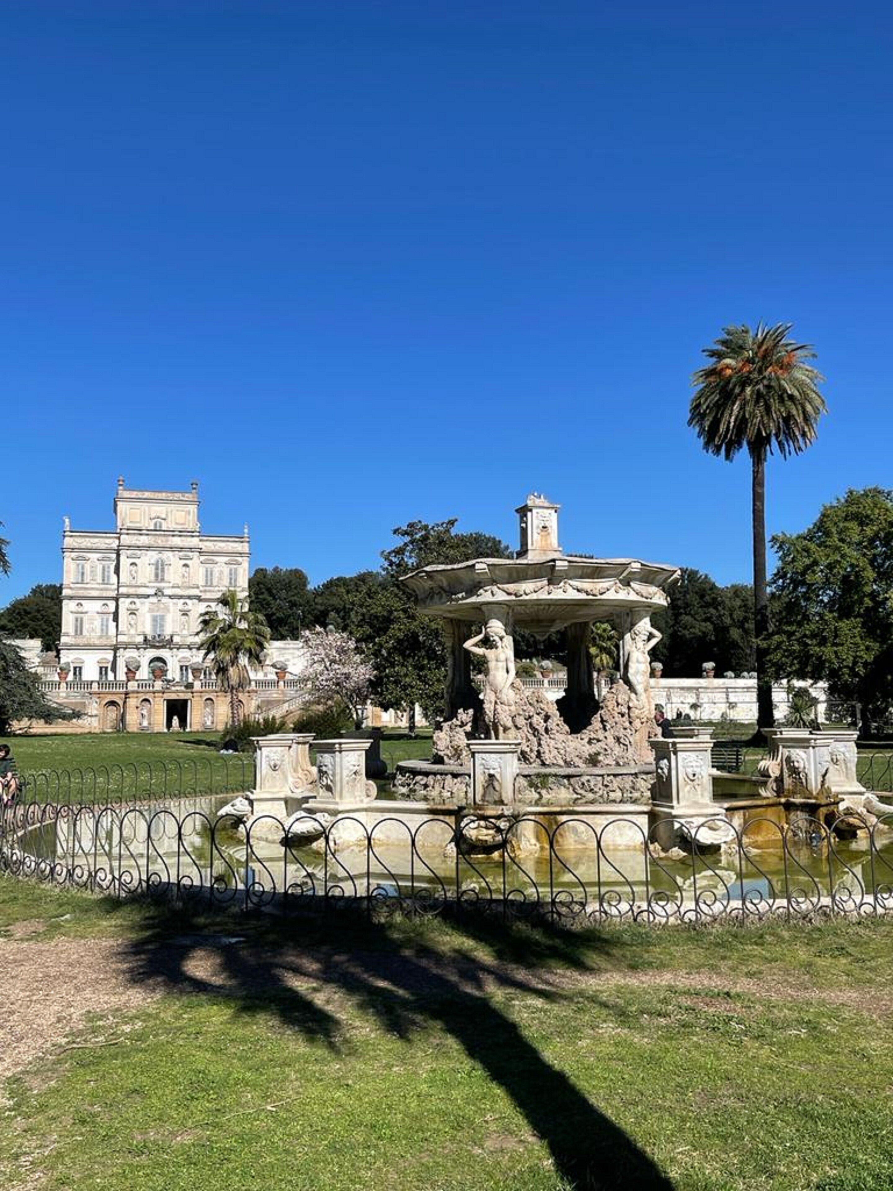 Una font clàssica amb estàtues, envoltada per una tanca de metall, a un parc amb un edifici gran al fons.