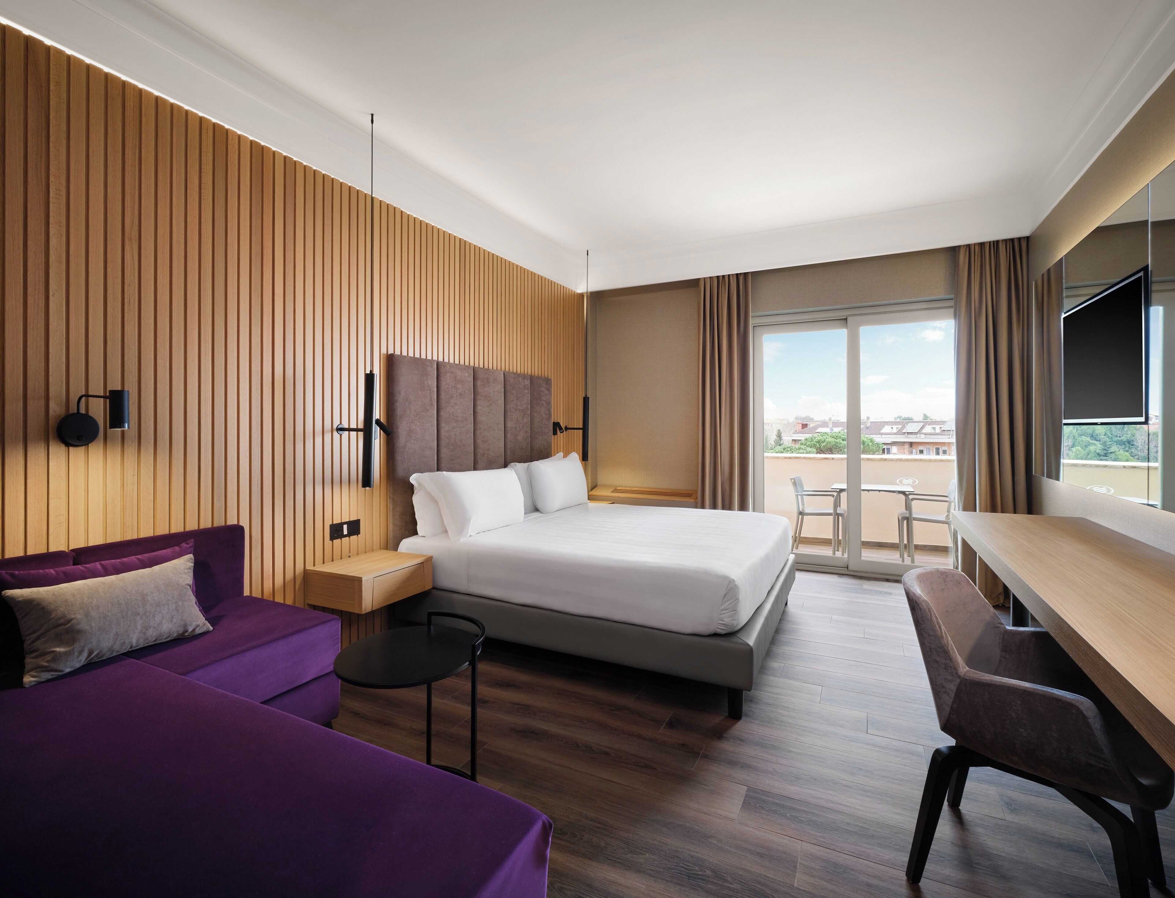 A modern hotel room with a large bed, a purple sofa, a small table, and a desk. The room has wooden paneling on the walls and a large window with a view.