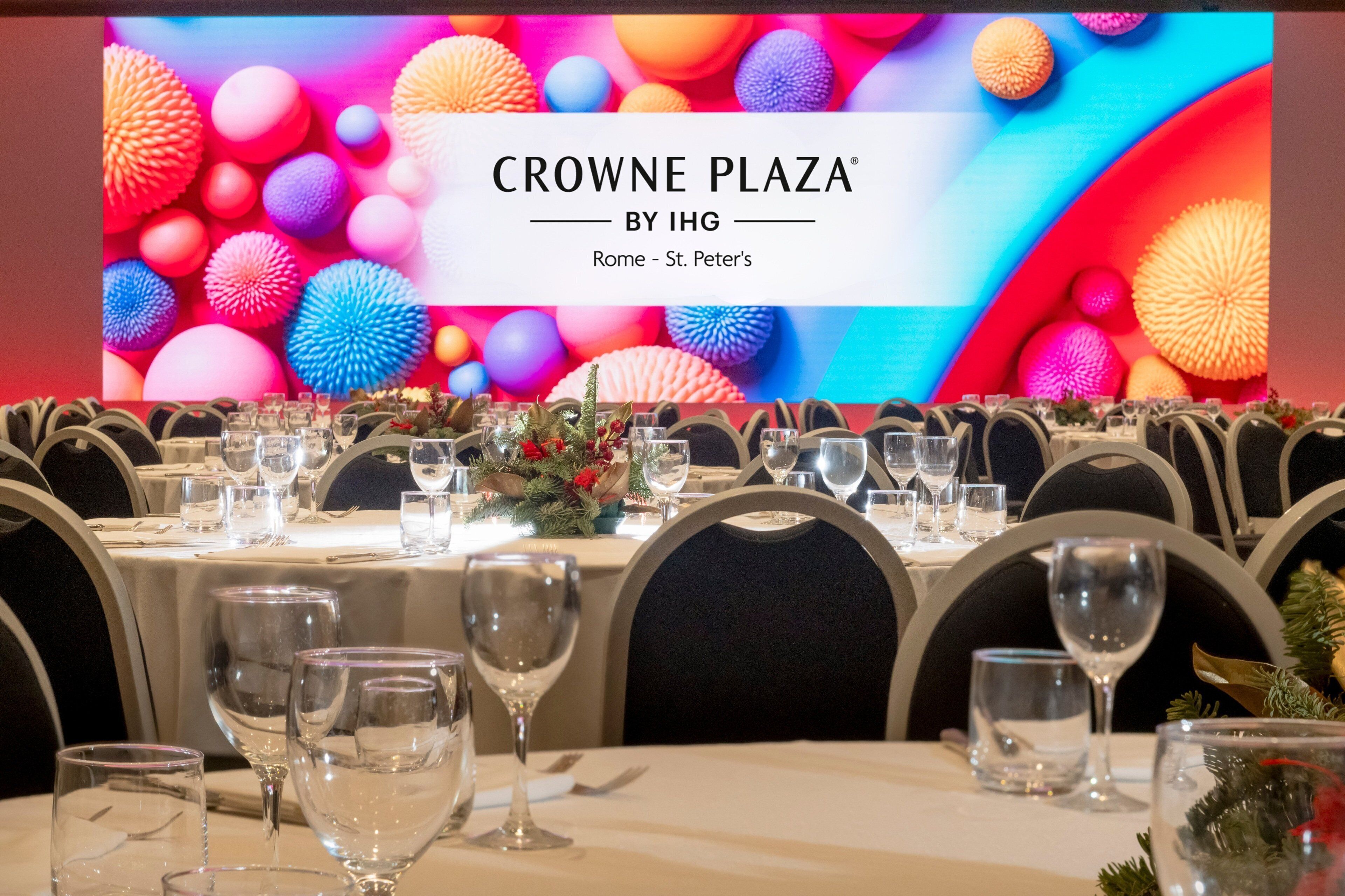 Ein Essbereich mit gedeckten Tischen und einem großen Bildschirm, der 'CROWNE PLAZA BY IHG Rom - Vatikan' anzeigt.