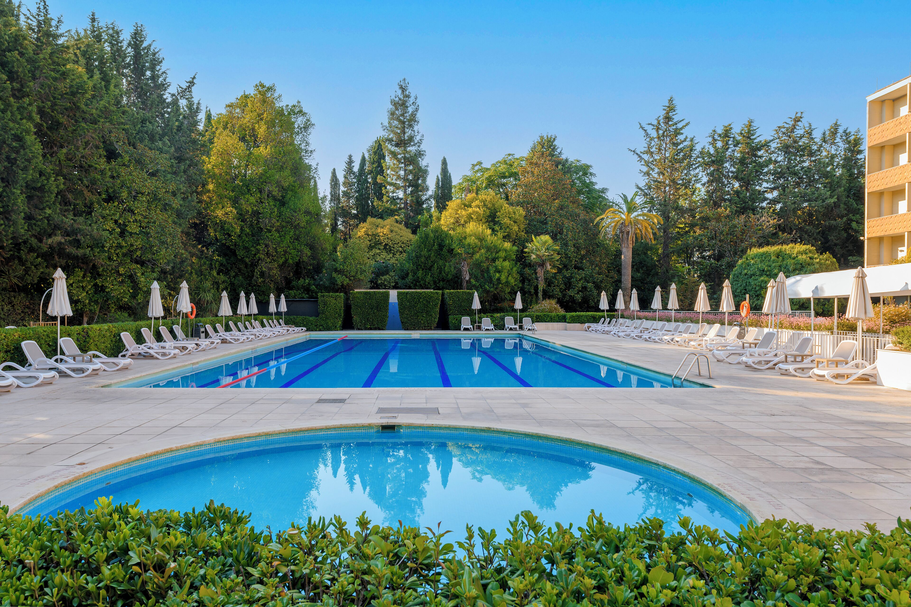 Una piscina con varias pistas, rodeada de sillas de descanso y sombrillas, con árboles y un edificio al fondo.