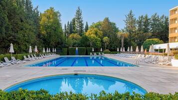 A swimming pool with multiple lanes, surrounded by lounge chairs and umbrellas, with trees and a building in the background.