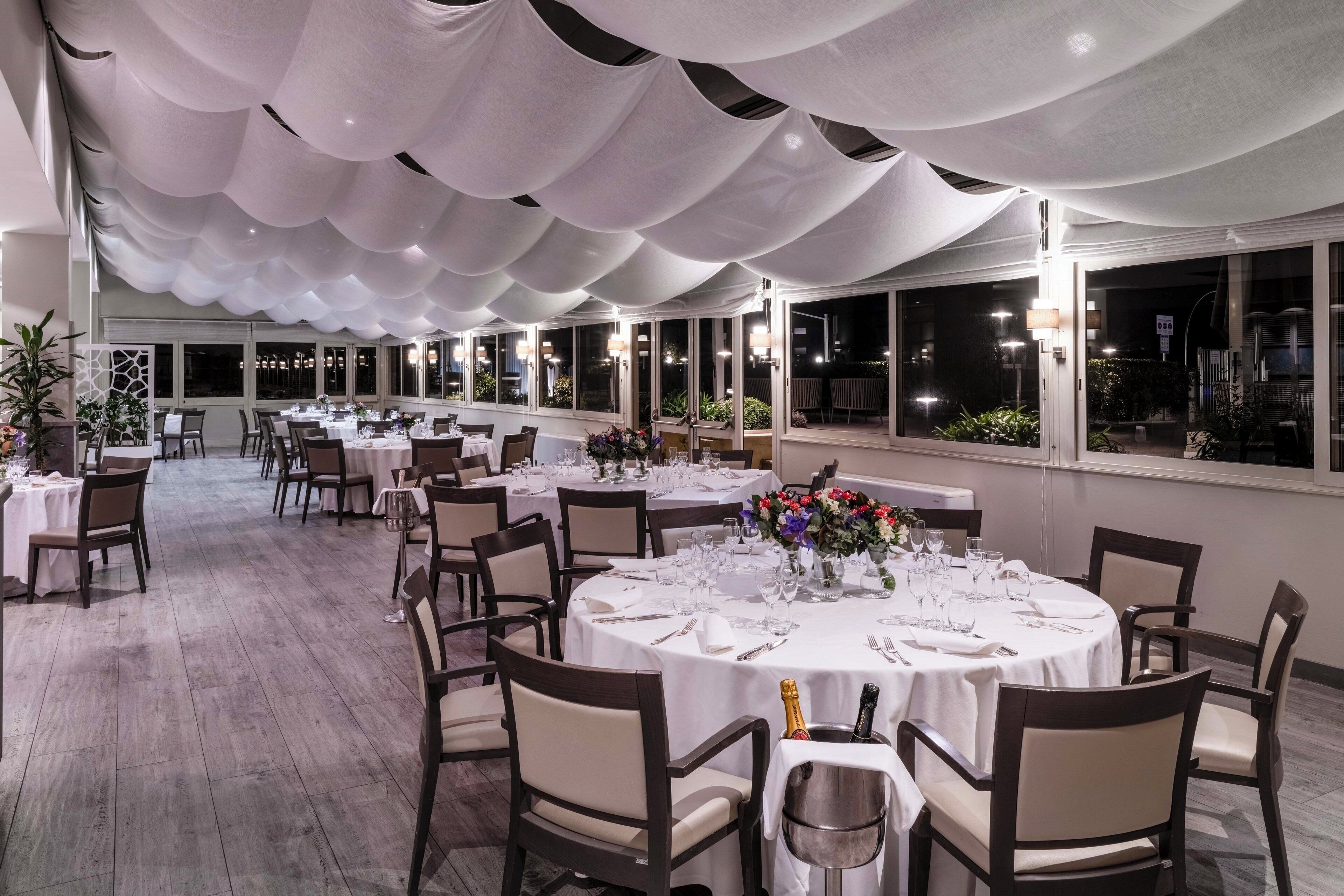 A spacious indoor dining area with round tables set for a meal, featuring white tablecloths and chairs.