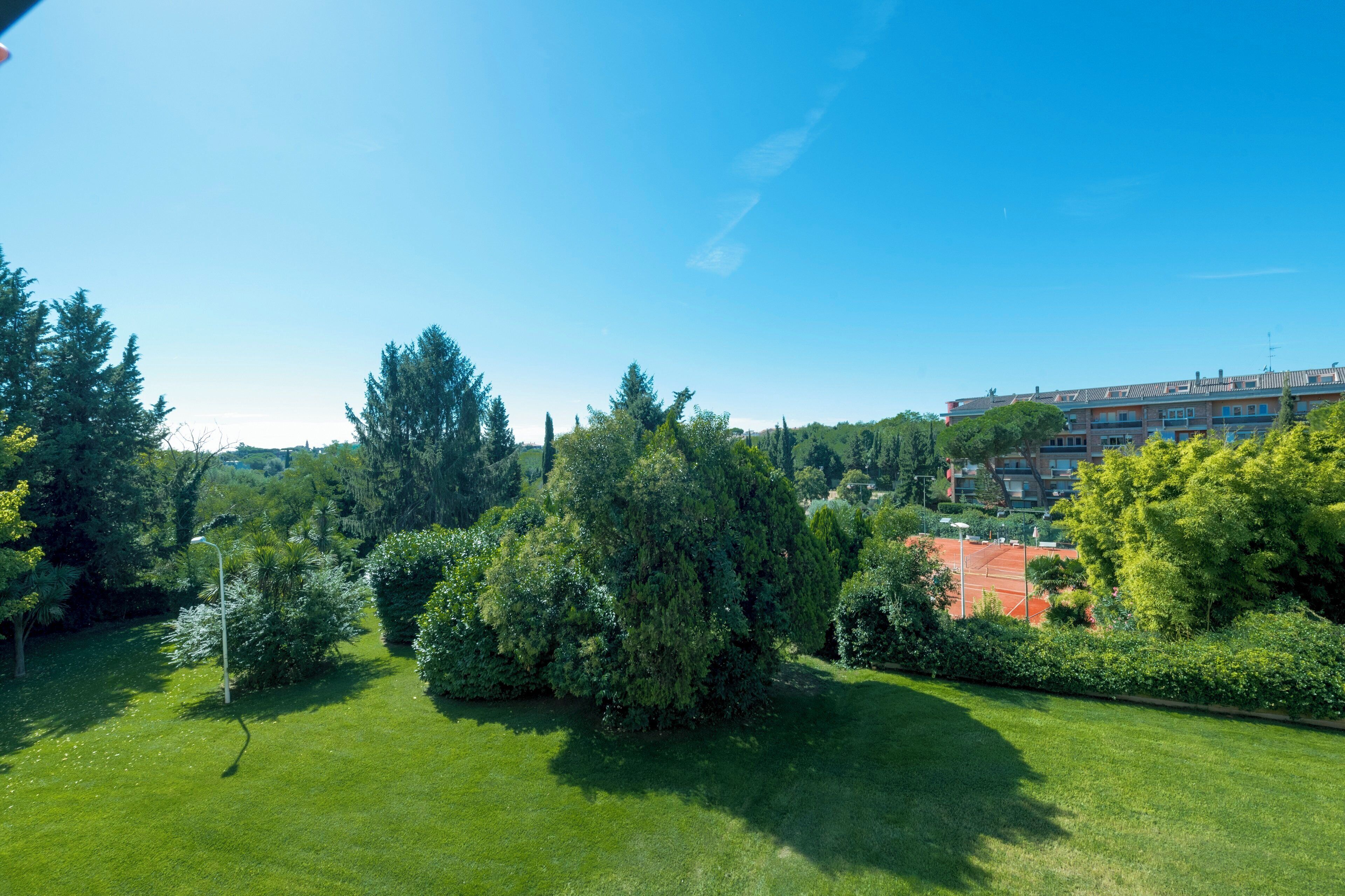 A well-maintained lawn with a tennis court, surrounded by dense trees and a multi-story building in the background.