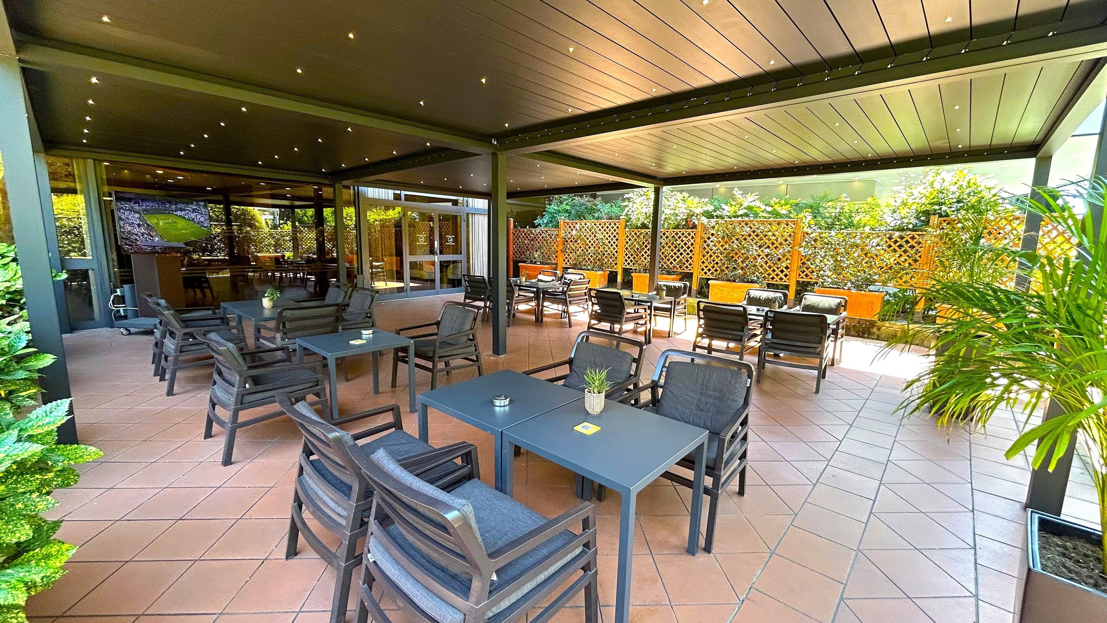 En udendørs terrasse med flere borde og stole, et overdækket område med tag, og udsigt til en have gennem vinduerne.