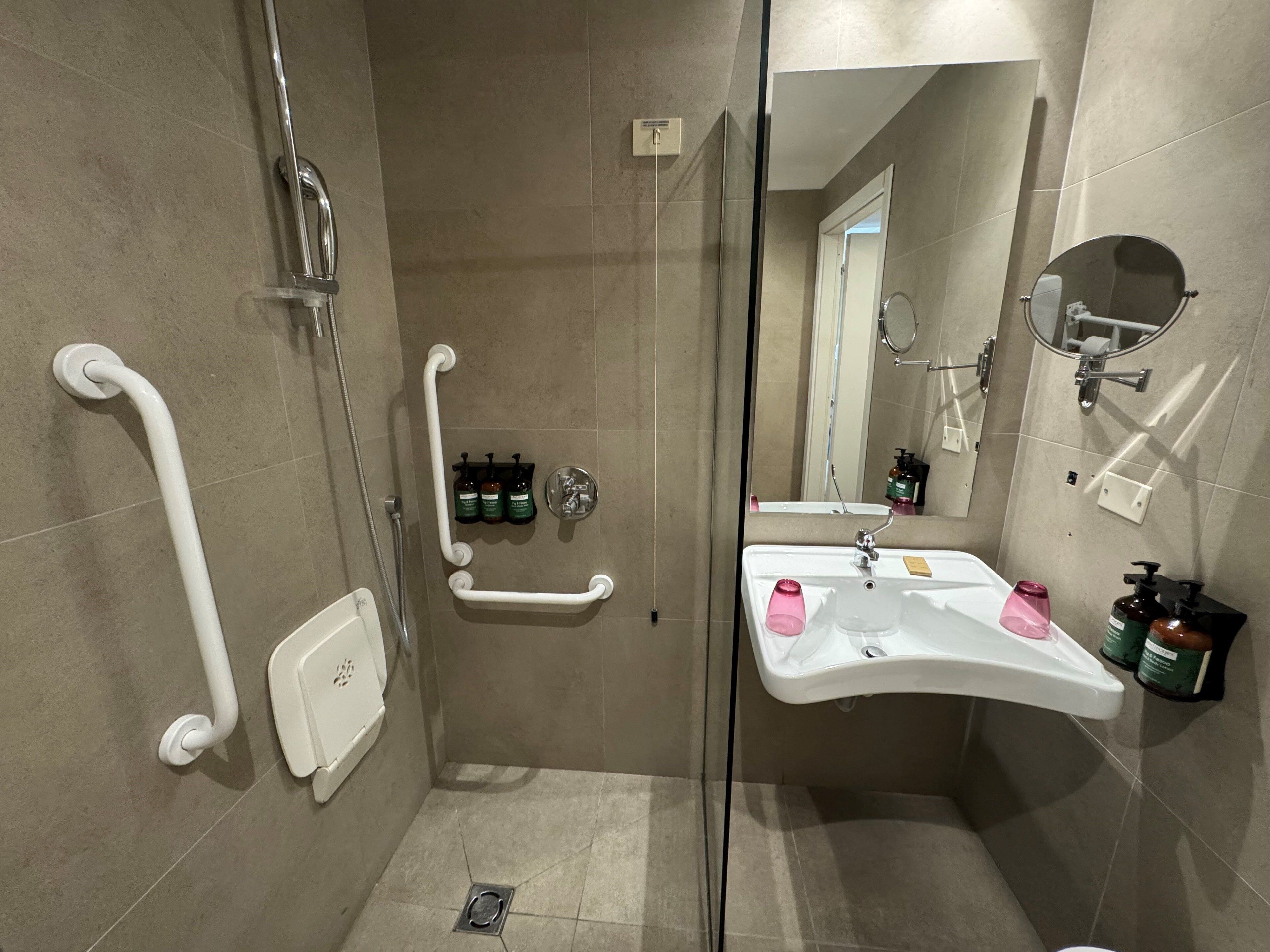 Uma casa de banho moderna com cabine de duche em vidro, lavatório branco com espelho e barra de apoio para acessibilidade.