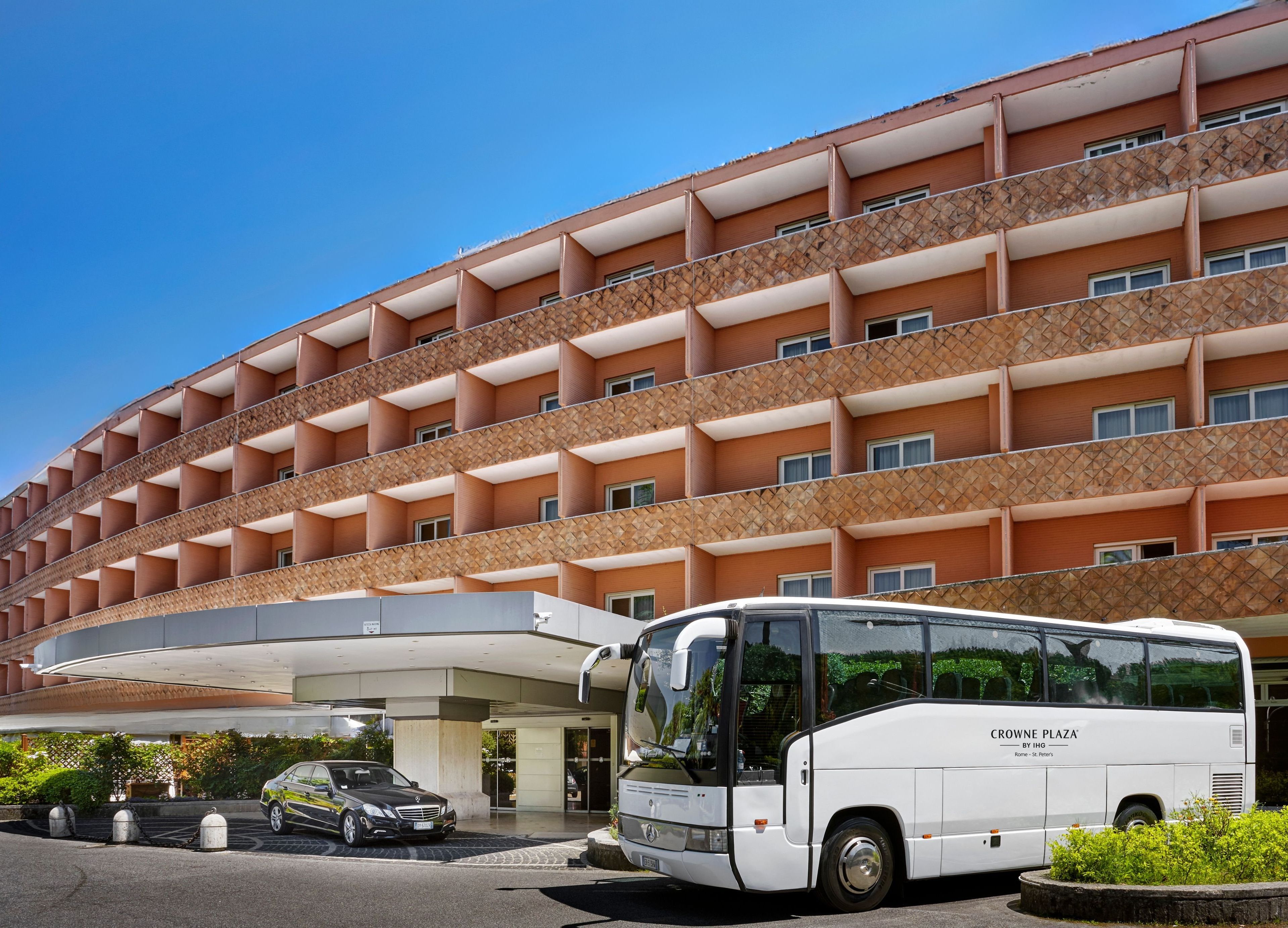 A multi-story hotel building with a curved facade, a white bus parked in front, and a black car in the parking area.