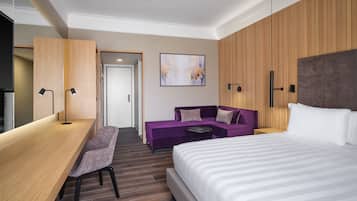 A modern hotel room with a large bed, a desk with a chair, a purple sofa, and a small table.