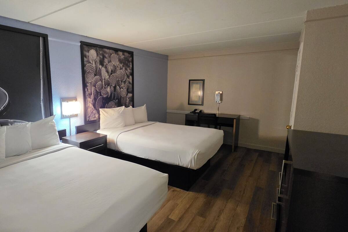 A hotel room with two beds, a desk, a chair, a lamp, and a guitar artwork on the wall.