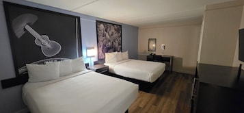 A hotel room with two beds, a desk, a chair, a lamp, and a guitar artwork on the wall.