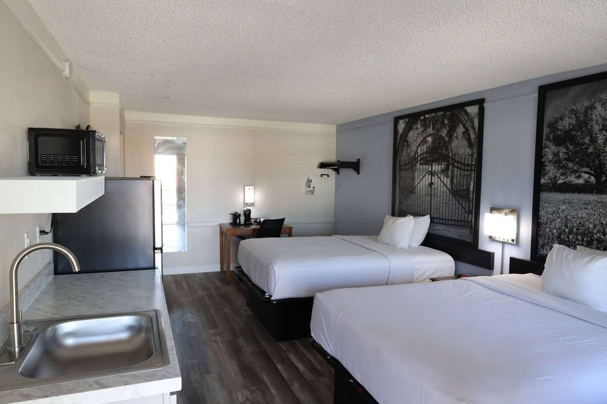 A hotel room with two beds, a small kitchenette, a sink, a microwave, and a painting on the wall.