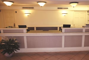 Un'area di accoglienza con un banco reception, due postazioni computer, una pianta in vaso e una porta.