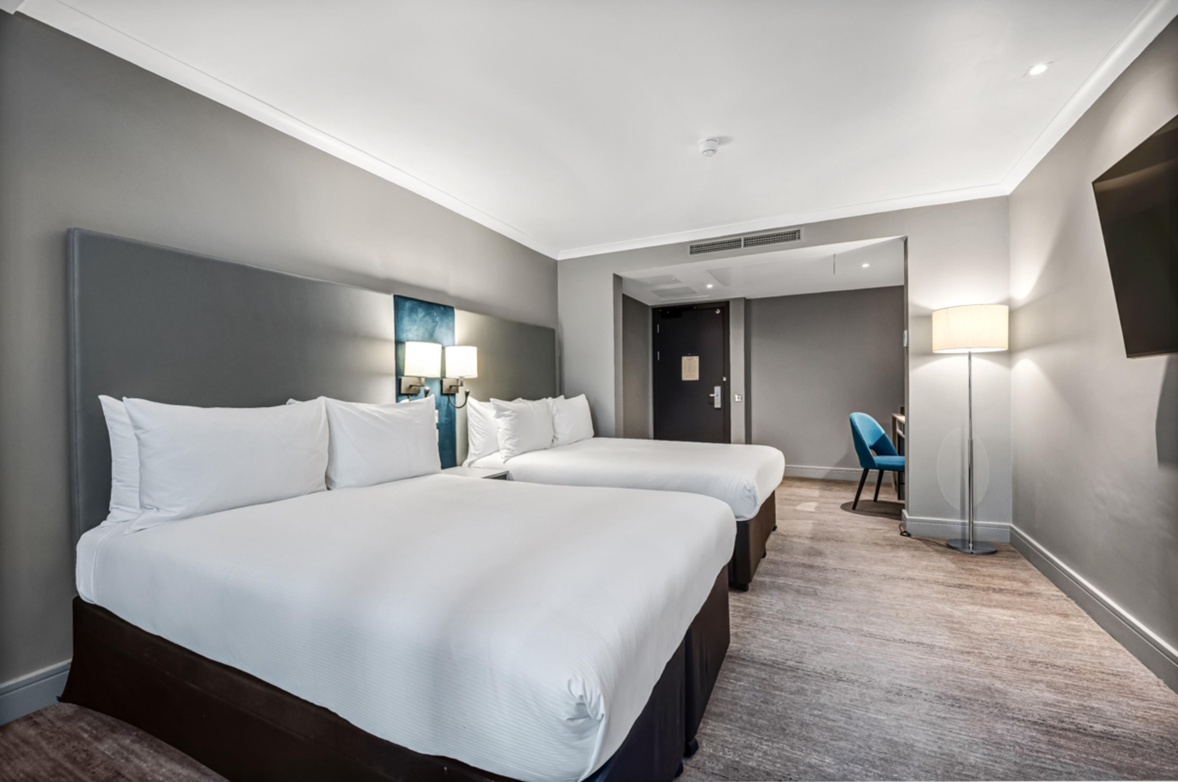A hotel room with two beds, a desk, a chair, a flat-screen TV, and a lamp.
