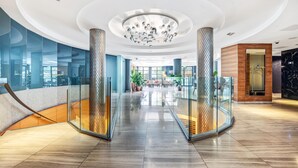 Un hall d’hôtel moderne avec une rambarde en verre, un lustre central et un comptoir d’accueil en bois.