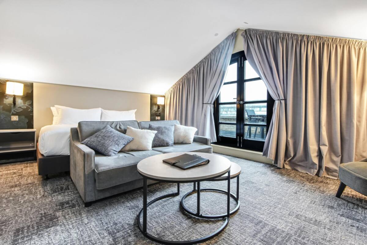 A hotel room with a bed, a sofa, a small round table, a chair, and a large window with curtains.