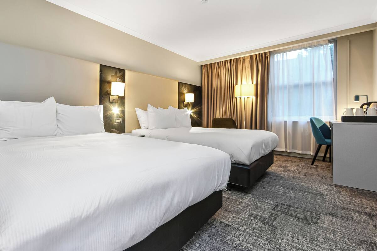 A hotel room with two beds, a desk, a chair, and a large window with curtains.