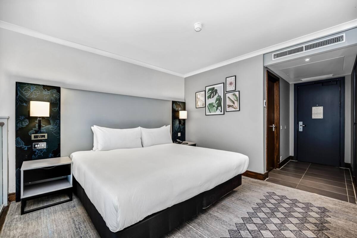 A hotel room with a large bed, bedside tables, a desk, a chair, a wall-mounted lamp, a framed picture, and a door.
