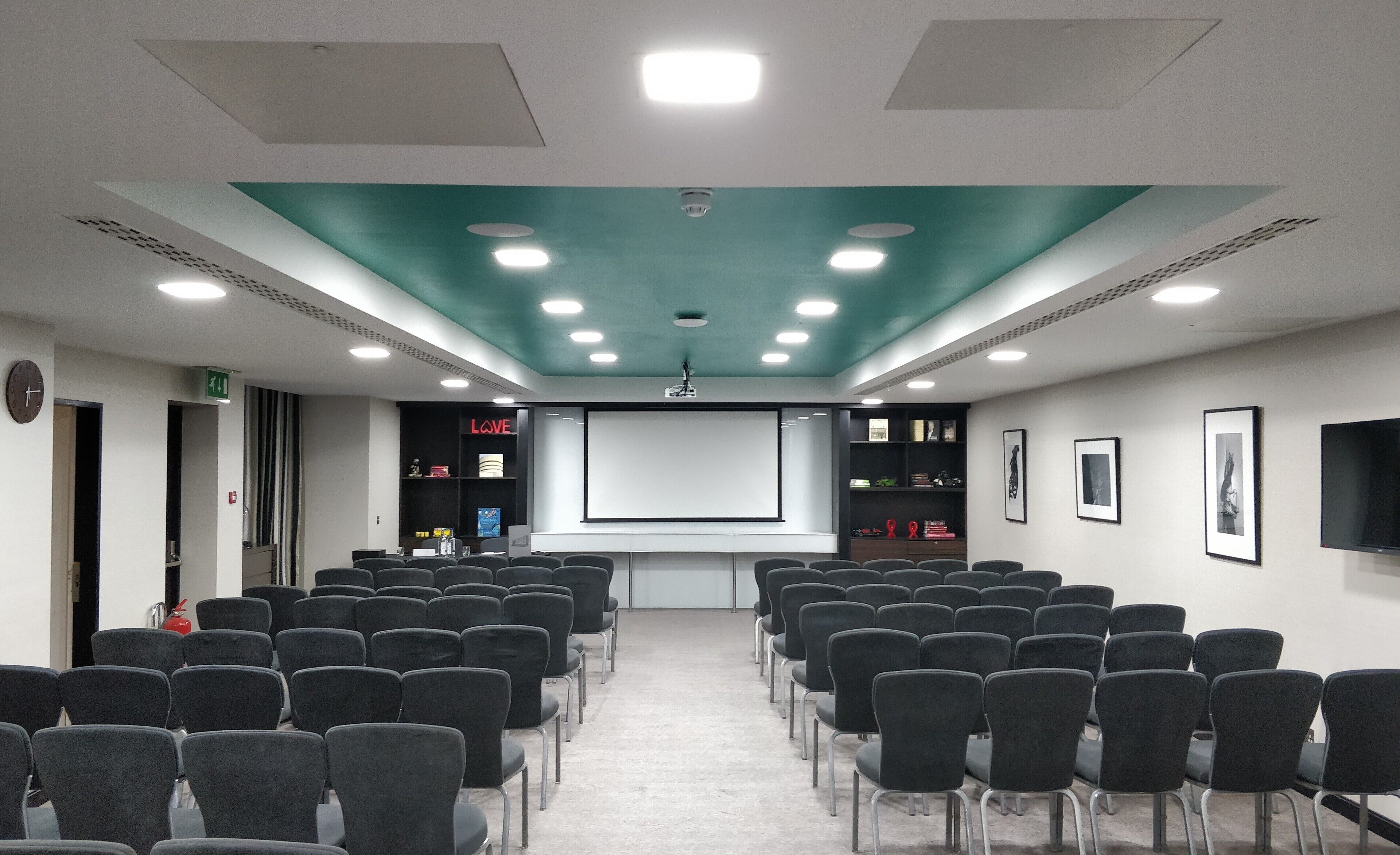A conference room with rows of chairs, a projector screen, a screen displaying 'LOVE', and a shelf with books and decorative items.